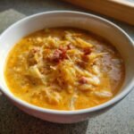 Close-up of a white bowl filled with rich, orange-hued Creamy Reuben Soup, topped with shredded cabbage and bits of bacon.