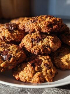 A stack of freshly baked, golden brown Crispy Chocolate Oat Cookies piled high on a white plate.