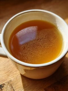 Close-up of a rustic mug filled with rich, golden Homemade Bone Broth resting on a sunlit wooden table.