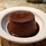 A perfectly molded, rich, dark chocolate pudding sitting in a pool of sauce in a white bowl.