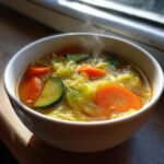 Close-up of a steaming white bowl filled with vibrant French Detox Soup featuring carrots, zucchini, and cabbage.