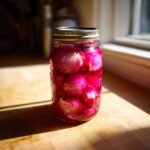 A glass jar filled with vibrant pink Gordon Ramsay Pickled Onions resting on a sunlit wooden counter.