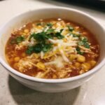 A close-up of a bowl of Green Chile Chicken Enchilada Soup topped with melted cheese, corn, and fresh cilantro.