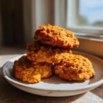 A stack of four golden-orange Healthy Pumpkin Oatmeal Cookies resting on a light ceramic plate near a window.