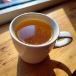 A close-up of a white mug filled with rich, golden Homemade Bone Broth sitting on a sunlit wooden surface.