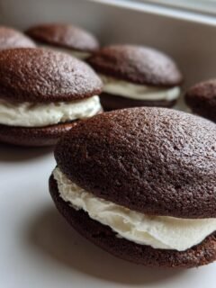 Several rich chocolate Gluten Free Whoopie Pies filled with thick white cream frosting sitting on a white surface.