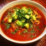 A close-up of a bowl of rich, red Instant Pot Chicken Enchilada Soup topped with diced avocado and fresh cilantro.