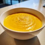 A close-up of a white bowl filled with bright orange Mango Gazpacho, drizzled with olive oil and pepper.