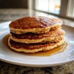 A close-up of three fluffy Oat Flour Pancakes stacked high and drizzled with maple syrup on a white plate.
