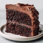 A close-up shot of a rich, dark slice of Paleo Chocolate Cake With Whipped Chocolate Frosting on a light plate.