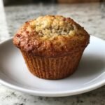 A freshly baked, golden-brown Protein Banana Muffin sitting on a small white plate.