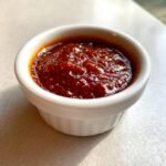 A small white ramekin filled with thick, dark red, glossy Shut Yo Mouth Sauce, showing texture and spices.