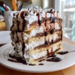 A tall slice of layered Homemade Ice Cream Cake featuring vanilla cake, chocolate mousse, and whipped cream, drizzled heavily with chocolate sauce.