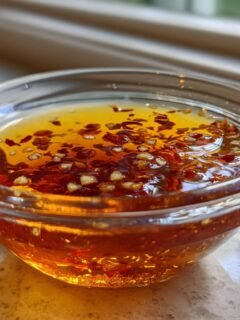 Close-up of a small glass bowl filled with golden Thai Dipping Sauce topped with red chili flakes and minced garlic.