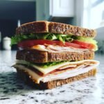 A tall Ultimate After The Pool Sandwich stacked high with turkey, cheese, tomato, and lettuce on dark wheat bread.