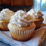 Three vanilla Dairy Free Cupcakes topped with swirls of fluffy white frosting resting on a white cloth.